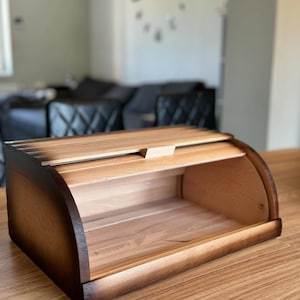 Puede incluir: Una panera de madera con tapa curva enrollable. La caja es de madera clara con bordes m&aacute;s oscuros y quemados. El interior es visible, mostrando la veta natural de la madera. La panera est&aacute; sobre una mesa de madera.
