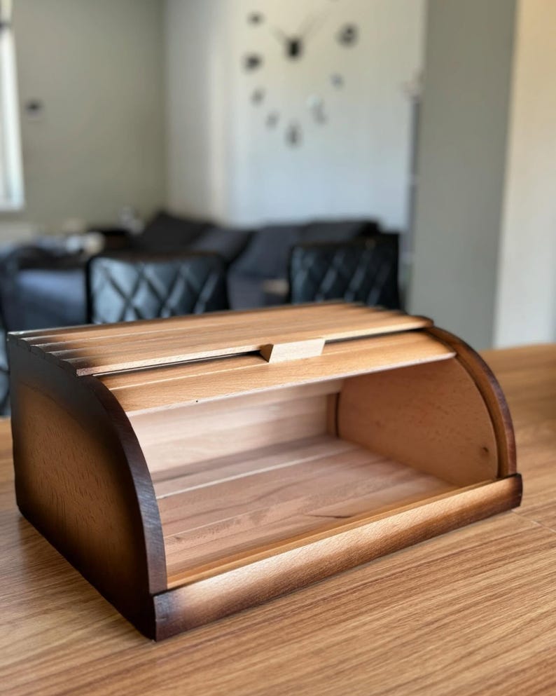 Puede incluir: Una panera de madera con tapa curva enrollable. La caja es de madera clara con bordes m&aacute;s oscuros y quemados. El interior es visible, mostrando la veta natural de la madera. La panera est&aacute; sobre una mesa de madera.