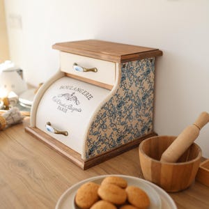 Puede incluir: Una panera de madera con frente color crema que incluye el texto BOULANGERIE, Cafe Chaud et Beignets, PARIS. Los laterales tienen un estampado floral azul y blanco, y cuenta con dos cajones con tiradores de lat&oacute;n y cer&aacute;mica azul.