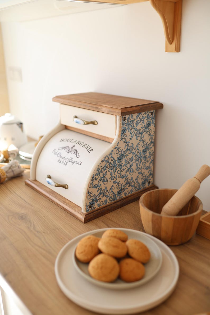 Puede incluir: Una panera de madera con frente color crema que incluye el texto BOULANGERIE, Cafe Chaud et Beignets, PARIS. Los laterales tienen un estampado floral azul y blanco, y cuenta con dos cajones con tiradores de lat&oacute;n y cer&aacute;mica azul.