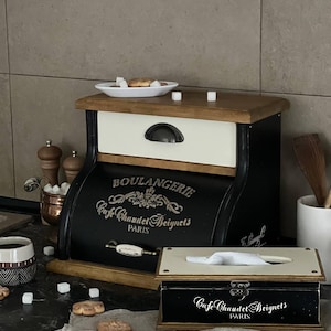 Puede incluir: Una panera y una caja de pa&ntilde;uelos negras y crema con letras doradas que dicen "BOULANGERIE Cafe Chaudet Beignets PARIS". La panera tiene una tapa de madera y un caj&oacute;n. La caja de pa&ntilde;uelos es rectangular con tapa abisagrada.