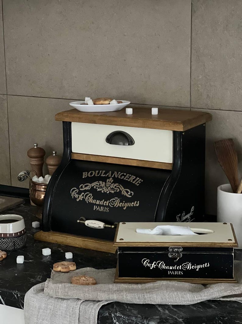 Puede incluir: Una panera y una caja de pa&ntilde;uelos negras y crema con letras doradas que dicen "BOULANGERIE Cafe Chaudet Beignets PARIS". La panera tiene una tapa de madera y un caj&oacute;n. La caja de pa&ntilde;uelos es rectangular con tapa abisagrada.
