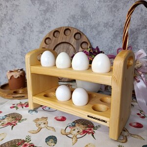 May include: A light-coloured wooden egg holder with two tiers and six circular indentations for storing eggs. The rack features a natural wood grain finish and the text BOOWOOD.RU is printed on the lower right side. Six white eggs are placed in the holder.