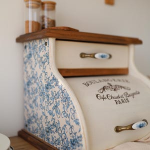 Puede incluir: Una panera de madera con puerta enrollable y un caj&oacute;n superior. Los laterales presentan un estampado floral azul y blanco. La puerta frontal tiene el texto BOULANGERIE, Cafe Chaud et Beignets, PARIS y un tirador de lat&oacute;n.