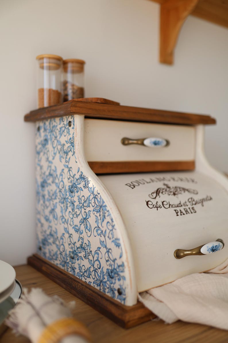 Puede incluir: Una panera de madera con puerta enrollable y un caj&oacute;n superior. Los laterales presentan un estampado floral azul y blanco. La puerta frontal tiene el texto BOULANGERIE, Cafe Chaud et Beignets, PARIS y un tirador de lat&oacute;n.