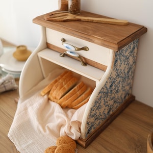 Puede incluir: Una panera de madera con frente color crema, tapa de madera natural y paneles laterales con estampado floral azul y blanco. Incluye un caj&oacute;n con dos tiradores de lat&oacute;n y cer&aacute;mica, conteniendo pan rebanado sobre un pa&ntilde;o claro y galletas cerca.