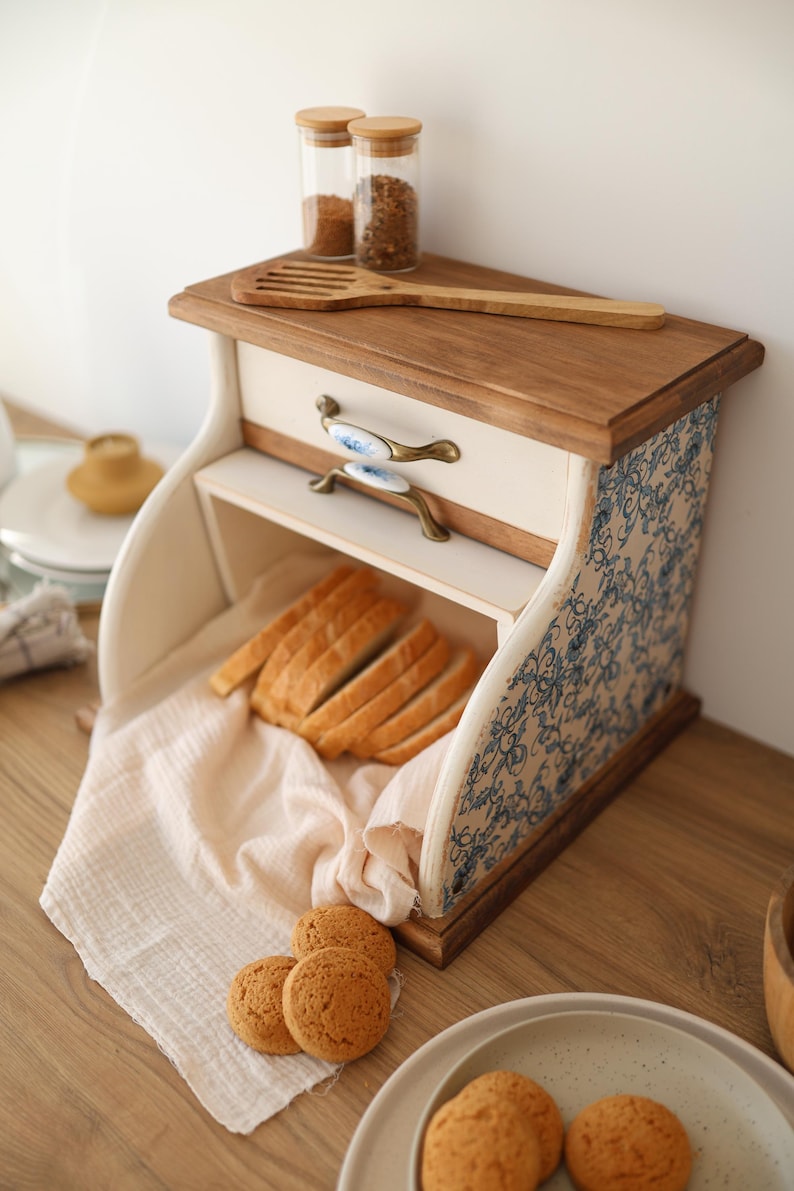 Puede incluir: Una panera de madera con frente color crema, tapa de madera natural y paneles laterales con estampado floral azul y blanco. Incluye un caj&oacute;n con dos tiradores de lat&oacute;n y cer&aacute;mica, conteniendo pan rebanado sobre un pa&ntilde;o claro y galletas cerca.