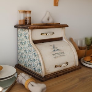 May include: A wooden bread box with a roll-top door featuring a blue windmill graphic and the text PATISSERIE DE PARIS. The sides are decorated with a blue floral pattern, and the unit includes a top drawer with a white and gold metal handle.