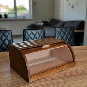 Puede incluir: Una panera de madera con tapa enrollable. La caja es de madera marr&oacute;n clara y oscura, con frente curvo y parte superior de listones. El interior es visible, mostrando madera de color claro.