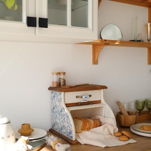 Puede incluir: Una panera de madera con un caj&oacute;n blanco con un patr&oacute;n floral azul en los laterales, un caj&oacute;n extra&iacute;ble con un tirador decorativo y un compartimento abierto que contiene pan rebanado sobre una servilleta de tela clara en una encimera.