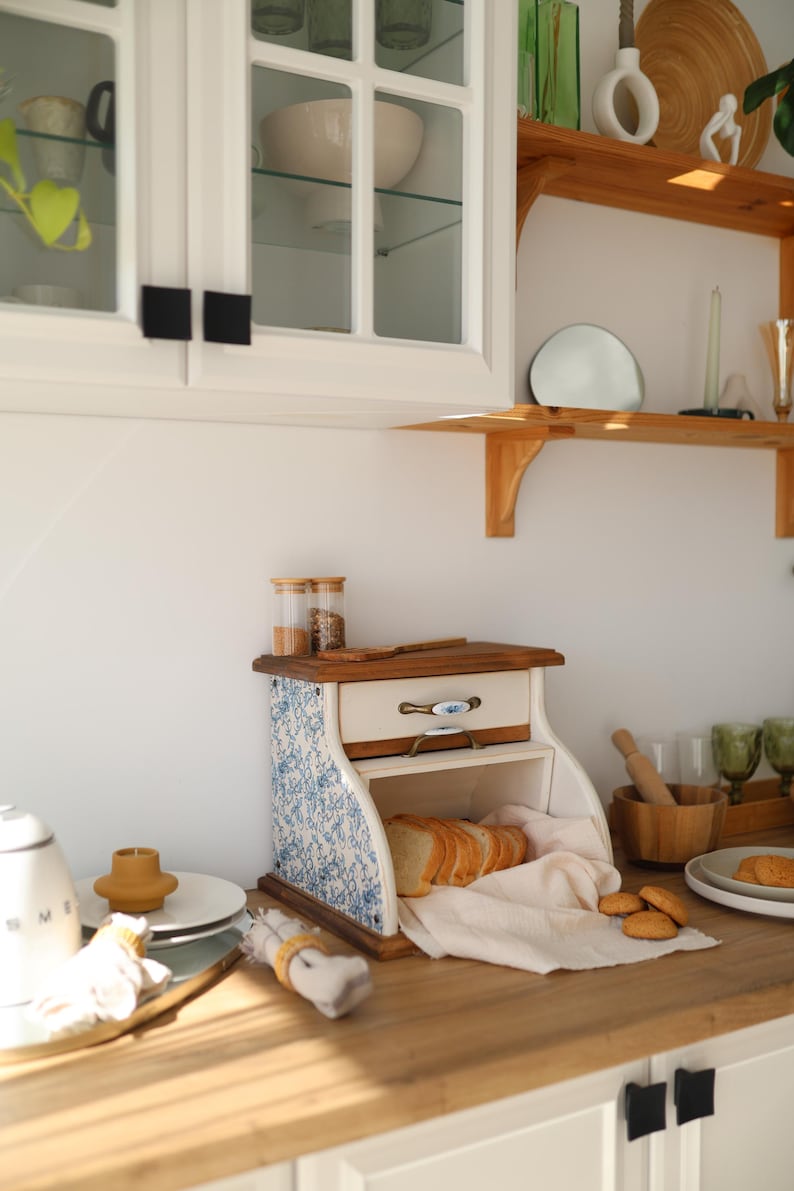 Puede incluir: Una panera de madera con un caj&oacute;n blanco con un patr&oacute;n floral azul en los laterales, un caj&oacute;n extra&iacute;ble con un tirador decorativo y un compartimento abierto que contiene pan rebanado sobre una servilleta de tela clara en una encimera.
