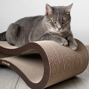 以下が含まれることがあります： 茶色とベージュの段ボール製の猫用爪とぎの上に横になっている灰色の猫。爪とぎは波の形をしていて、下部に「Cat Joy」の文字が書かれている。