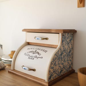 Puede incluir: Una panera de madera color crema con puerta enrollable y un caj&oacute;n superior. El frente presenta el texto negro BOULANGERIE Cafe Chaud et Beignets PARIS. Los lados tienen un patr&oacute;n floral azul y los tiradores de lat&oacute;n tienen detalles florales azules.