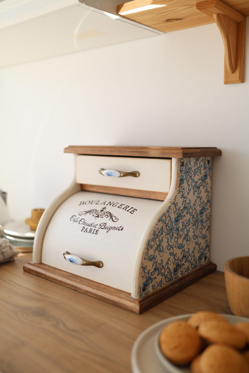 Puede incluir: Una panera de madera color crema con puerta enrollable y un caj&oacute;n superior. El frente presenta el texto negro BOULANGERIE Cafe Chaud et Beignets PARIS. Los lados tienen un patr&oacute;n floral azul y los tiradores de lat&oacute;n tienen detalles florales azules.