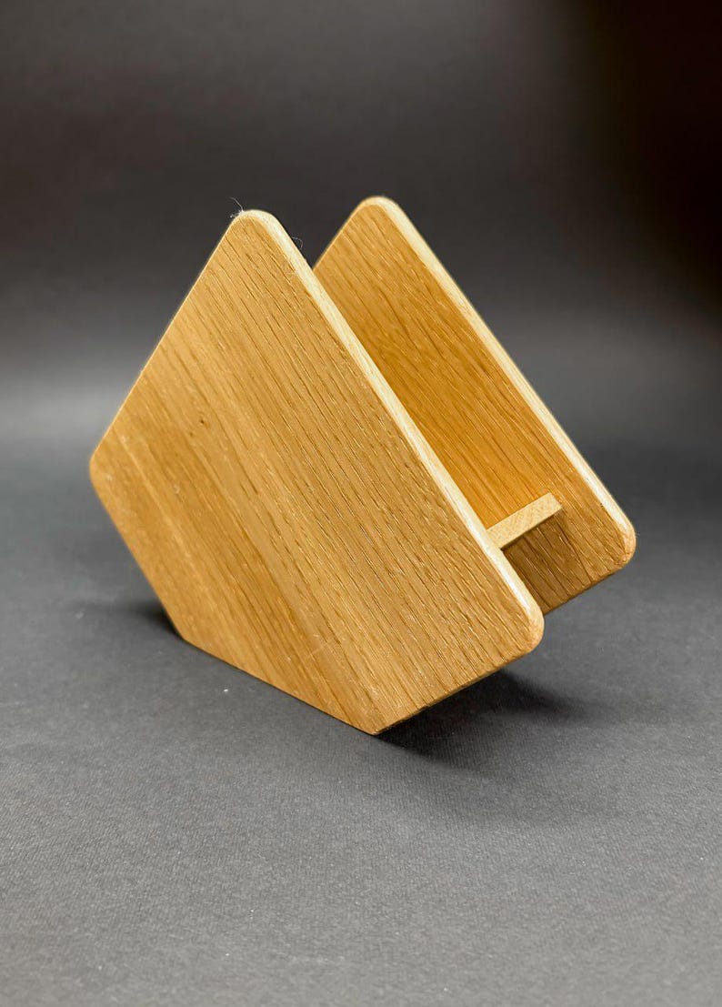 Puede incluir: Un servilletero de madera con un acabado de vetas naturales. El soporte est&aacute; construido con dos piezas de madera anguladas unidas en la base, creando una forma triangular. El soporte est&aacute; dise&ntilde;ado para sujetar servilletas en posici&oacute;n vertical.