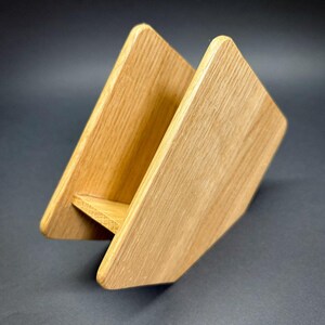 Puede incluir: Un servilletero de madera con acabado natural. El soporte est&aacute; hecho de dos piezas de madera anguladas unidas en la base, creando una forma triangular. La veta de la madera es visible y los bordes son redondeados.