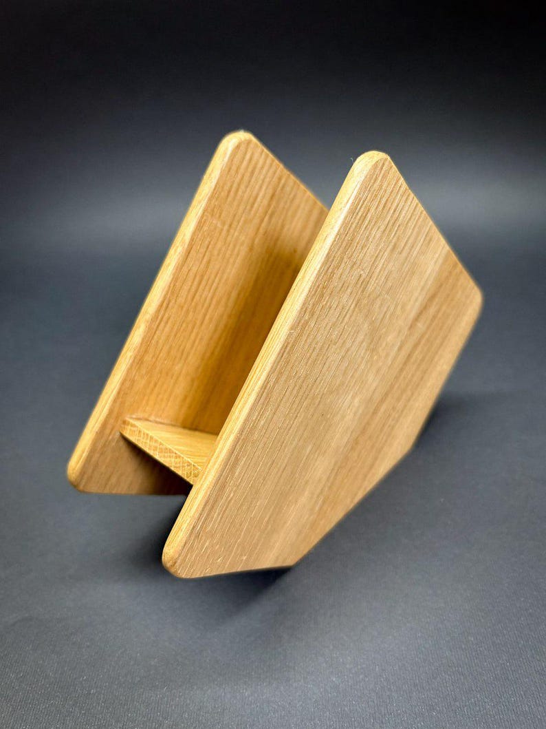 Puede incluir: Un servilletero de madera con acabado natural. El soporte est&aacute; hecho de dos piezas de madera anguladas unidas en la base, creando una forma triangular. La veta de la madera es visible y los bordes son redondeados.