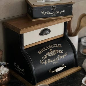 Puede incluir: Un juego de cajas de almacenamiento de madera de dos niveles, en negro y crema. La caja m&aacute;s grande presenta el texto "BOULANGERIE Cafe Chaudet Beignets PARIS". Una caja m&aacute;s peque&ntilde;a en la parte superior tiene el mismo texto. El conjunto tiene una est&eacute;tica vintage.