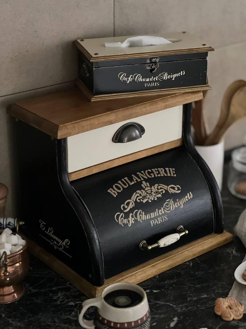 Puede incluir: Un juego de cajas de almacenamiento de madera de dos niveles, en negro y crema. La caja m&aacute;s grande presenta el texto "BOULANGERIE Cafe Chaudet Beignets PARIS". Una caja m&aacute;s peque&ntilde;a en la parte superior tiene el mismo texto. El conjunto tiene una est&eacute;tica vintage.