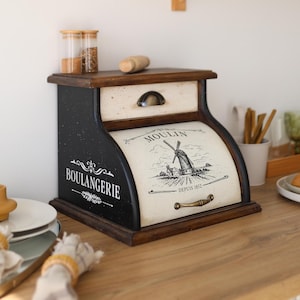 May include: A wooden bread box with a black frame and a cream-colored roll-top door. The door features a black graphic of a windmill and the text "MOULIN DEPUIS 1852". The side is labeled "BOULANGERIE" in white text. It includes a small top drawer.