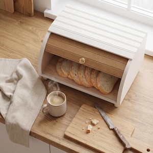 Puede incluir: Una panera blanca con puerta corredera de madera y pomo dorado, que contiene varias rebanadas de pan. Al lado hay una taza beige con café humeante, una tabla de cortar de madera con migas de pan y un cuchillo de sierra sobre una superficie.