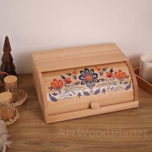 Op de afbeelding: Een houten broodtrommel met een roldeksel. De voorkant heeft een bloemenontwerp in oranje, blauw en wit. De trommel heeft een kleine houten knop en is gemaakt van lichtgekleurd hout. De tekst "ArtWoodPlanet" is zichtbaar.