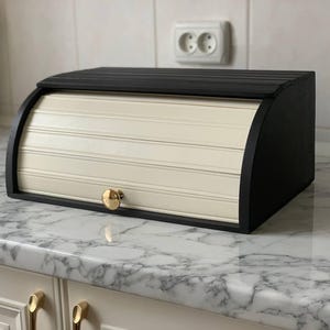 May include: A black and cream-colored wooden bread box with a roll-top door. The box has a curved top and a gold-colored knob. The bread box is sitting on a marble countertop.
