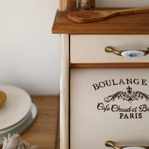 Puede incluir: Una panera de madera con un panel frontal de color crema que presenta el texto marr&oacute;n BOULANGERIE y Cafe Chaud et Be PARIS. La unidad tiene un caj&oacute;n superior y una puerta abatible, ambos con tiradores de lat&oacute;n con inserciones de cer&aacute;mica azul.