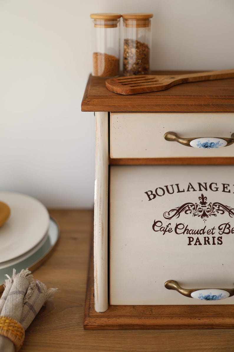 Puede incluir: Una panera de madera con un panel frontal de color crema que presenta el texto marr&oacute;n BOULANGERIE y Cafe Chaud et Be PARIS. La unidad tiene un caj&oacute;n superior y una puerta abatible, ambos con tiradores de lat&oacute;n con inserciones de cer&aacute;mica azul.