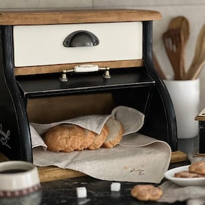 Puede incluir: Una panera r&uacute;stica de madera con marco negro, caj&oacute;n crema y tapa de madera natural. La panera abierta revela rebanadas de pan fresco sobre un pa&ntilde;o de lino. Una taza de caf&eacute; y galletas est&aacute;n en primer plano.