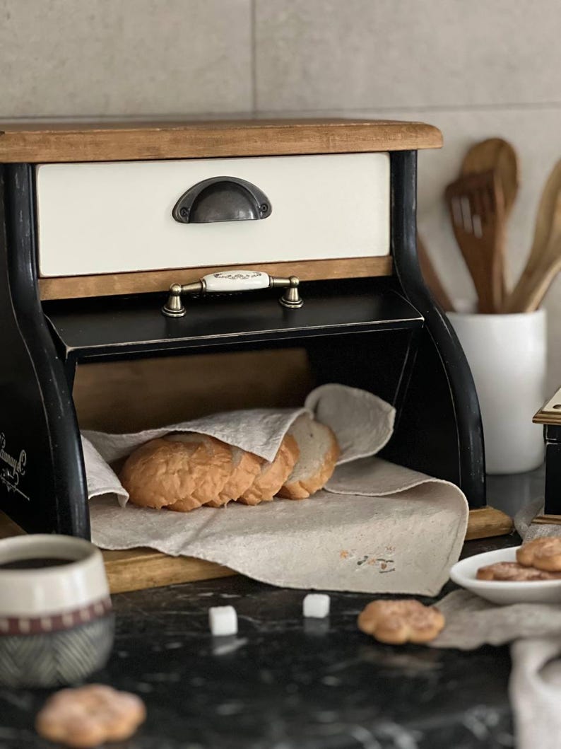 Puede incluir: Una panera r&uacute;stica de madera con marco negro, caj&oacute;n crema y tapa de madera natural. La panera abierta revela rebanadas de pan fresco sobre un pa&ntilde;o de lino. Una taza de caf&eacute; y galletas est&aacute;n en primer plano.