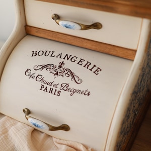 Puede incluir: Una panera de madera color crema con puerta enrollable y un caj&oacute;n superior. El frente presenta el texto marr&oacute;n BOULANGERIE, Cafe Chaud et Beignets, PARIS y un adorno decorativo. Tiene dos tiradores de lat&oacute;n con inserciones de cer&aacute;mica blanca.