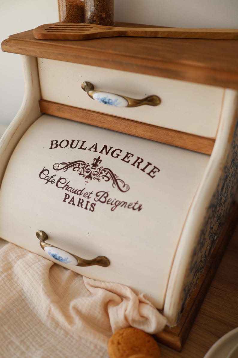 Puede incluir: Una panera de madera color crema con puerta enrollable y un caj&oacute;n superior. El frente presenta el texto marr&oacute;n BOULANGERIE, Cafe Chaud et Beignets, PARIS y un adorno decorativo. Tiene dos tiradores de lat&oacute;n con inserciones de cer&aacute;mica blanca.