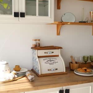Puede incluir: Una panera de madera con frente color crema que incluye el texto BOULANGERIE Cafe Chaud et Beignets PARIS. Los laterales tienen un estampado floral azul y blanco, e incluye dos cajones con tiradores de metal y una tapa superior plana.