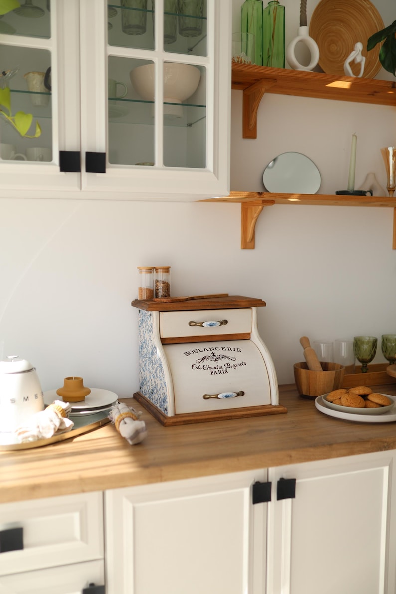 Puede incluir: Una panera de madera con frente color crema que incluye el texto BOULANGERIE Cafe Chaud et Beignets PARIS. Los laterales tienen un estampado floral azul y blanco, e incluye dos cajones con tiradores de metal y una tapa superior plana.