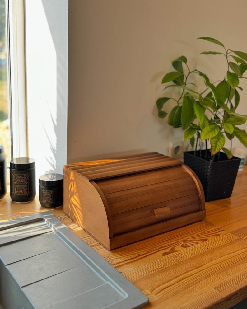 Puede incluir: Una panera de madera con tapa enrollable sobre una encimera de madera. La panera es de color marr&oacute;n c&aacute;lido y tiene un frente curvo. Dos botes negros con tapas est&aacute;n a la izquierda de la panera. Una peque&ntilde;a planta en maceta est&aacute; a la derecha.