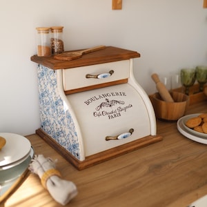 May include: A wooden bread box with a roll-top door featuring the text BOULANGERIE Cafe Chaud et Beignets PARIS. The sides have a blue and white floral pattern, and the unit includes a top drawer with a ceramic handle and a wooden top surface.
