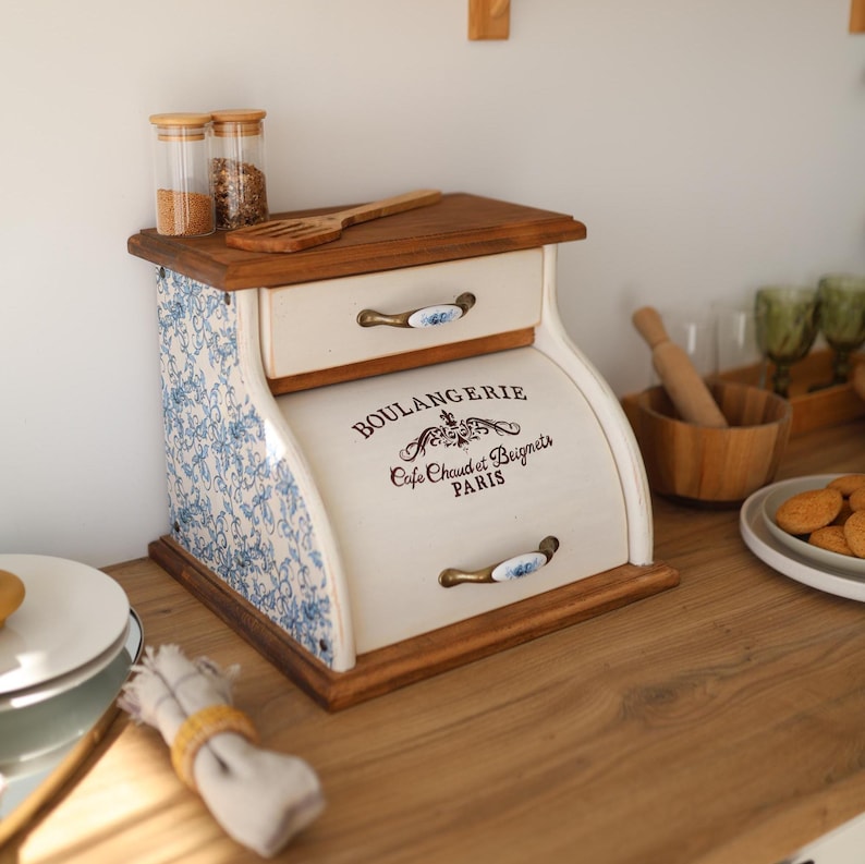Puede incluir: Una panera de madera con puerta enrollable que presenta el texto BOULANGERIE Cafe Chaud et Beignets PARIS. Los laterales tienen un patr&oacute;n floral azul y blanco, e incluye un caj&oacute;n superior con tirador de cer&aacute;mica y una superficie superior de madera.