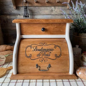 May include: A wooden bread bin with a roll-top door featuring the black text Boulangerie de Paris. The unit has a natural wood finish, white side supports, a top drawer with a black knob, and a decorative metal handle on the front door.