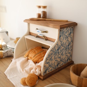 May include: A wooden bread box with a cream-colored finish and blue floral patterned side panels. It features a top drawer with two brass-toned handles, a roll-top opening containing sliced bread on a cloth, and a flat wooden top surface.