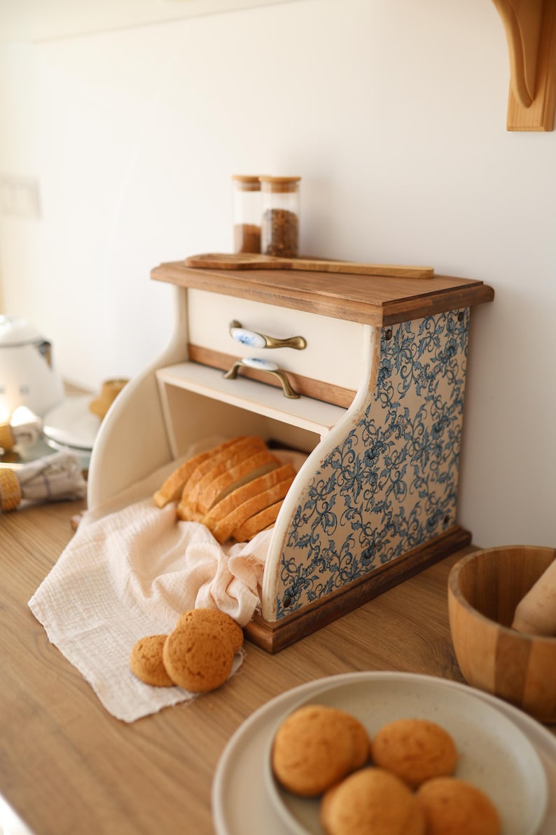 Puede incluir: Una panera de madera con acabado color crema y paneles laterales con estampado floral azul. Cuenta con un caj&oacute;n superior con dos tiradores de tono lat&oacute;n, una apertura enrollable con pan rebanado sobre un pa&ntilde;o y una superficie superior plana.