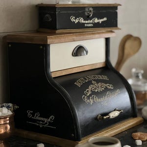 Puede incluir: Una panera negra y crema con el texto "BOULANGERIE Cafe Chandet Beignets PARIS" en letras doradas. La panera tiene un caj&oacute;n con un tirador de metal y una peque&ntilde;a caja de madera encima. La panera es de madera.