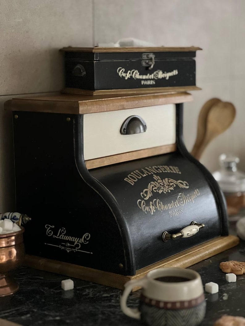 Puede incluir: Una panera negra y crema con el texto "BOULANGERIE Cafe Chandet Beignets PARIS" en letras doradas. La panera tiene un caj&oacute;n con un tirador de metal y una peque&ntilde;a caja de madera encima. La panera es de madera.