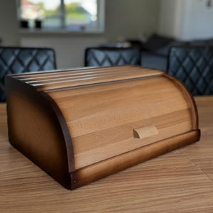 May include: A wooden bread bin with a roll-top lid. The bin is made of light-coloured wood with darker brown accents on the edges. It has a small wooden handle on the front and is sitting on a wooden table.