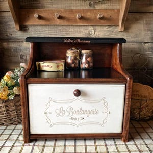 Puede incluir: Una panera de madera con acabado marrón oscuro y un cajón frontal blanco con el texto La Boulangerie Bakery. El estante superior es negro con el texto La Vie Parisienne y contiene dos pequeños frascos de vidrio llenos de nueces y una caja.