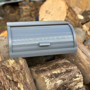 Könnte beinhalten: Eine graue Brotdose aus Holz mit Rolldeckel und kleinem Griff. Die Brotdose ist rechteckig und liegt auf einem Stapel Holzscheite. Die Holzscheite sind unterschiedlich groß und haben eine natürliche Holzmaserung.