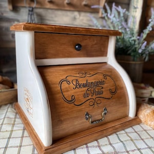 Könnte beinhalten: Ein Brotkasten aus Holz mit Rolltür und einer oberen Schublade. Die Vorderseite zeigt den schwarzen Schriftzug "Boulangerie de Paris" mit dekorativen Schnörkeln. Das Gehäuse hat eine naturfarbene Holzoberfläche, kontrastiert durch weiß lackierte Seitenteile und einen kleinen schwarzen Schubladenknopf.