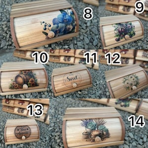 Op de afbeelding: Houten broodtrommels met diverse decoratieve ontwerpen. E&eacute;n doos heeft blauwe orchidee&euml;n, een andere zonnebloemen en een vat, en andere tonen bloemstukken, bessen en tekst. Elke doos heeft een gebogen bovenkant en een houten knop.