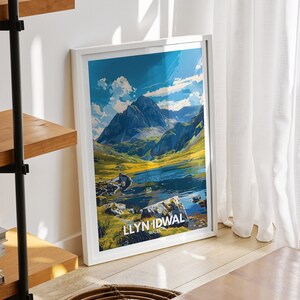 May include: A framed poster with a vintage travel design featuring a mountain lake and the text "LLYN IDWAL WALES". The poster is hanging on a white wall in a room with a wooden shelf and a wicker stool.