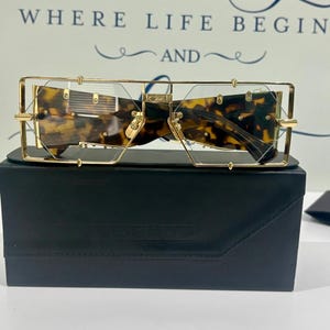 May include: Gold-tone metal frame sunglasses featuring a rectangular, geometric design with tortoiseshell-patterned lenses. The glasses are positioned atop a black rectangular case. The frame includes thin metal bars and small screw details.